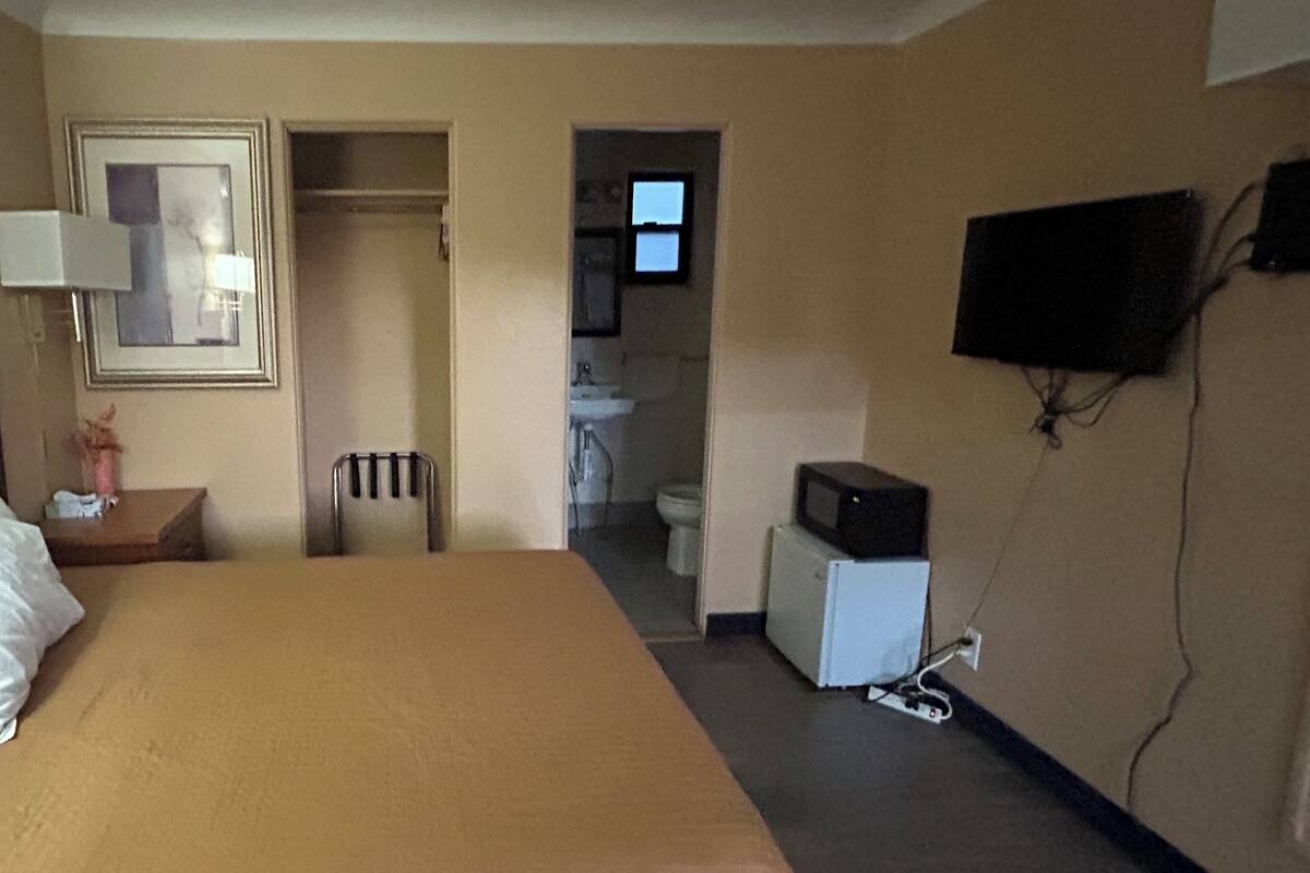 A hotel room with a bed, a desk, a chair, a television mounted on the wall, and a small kitchenette area.