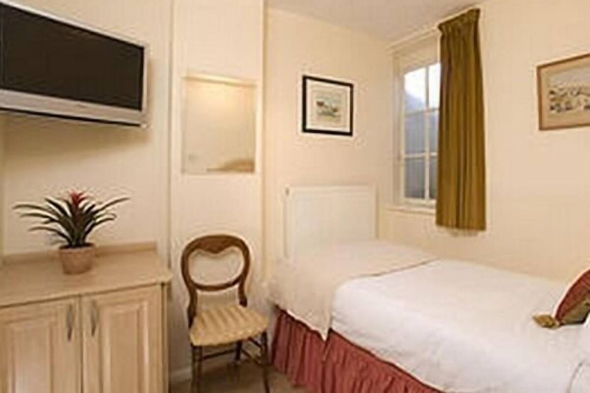 A small, single-bed room with a television, a chair, a radiator, and a window with curtains.