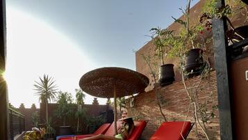 A sunlit outdoor lounge area with red cushioned chairs and a large umbrella, surrounded by potted plants and a brick wall.