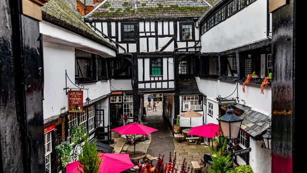 A narrow alleyway with traditional half-timbered buildings, outdoor seating with umbrellas, and a visible hotel reception. - The New Inn by Roomsbooked (Gloucester)