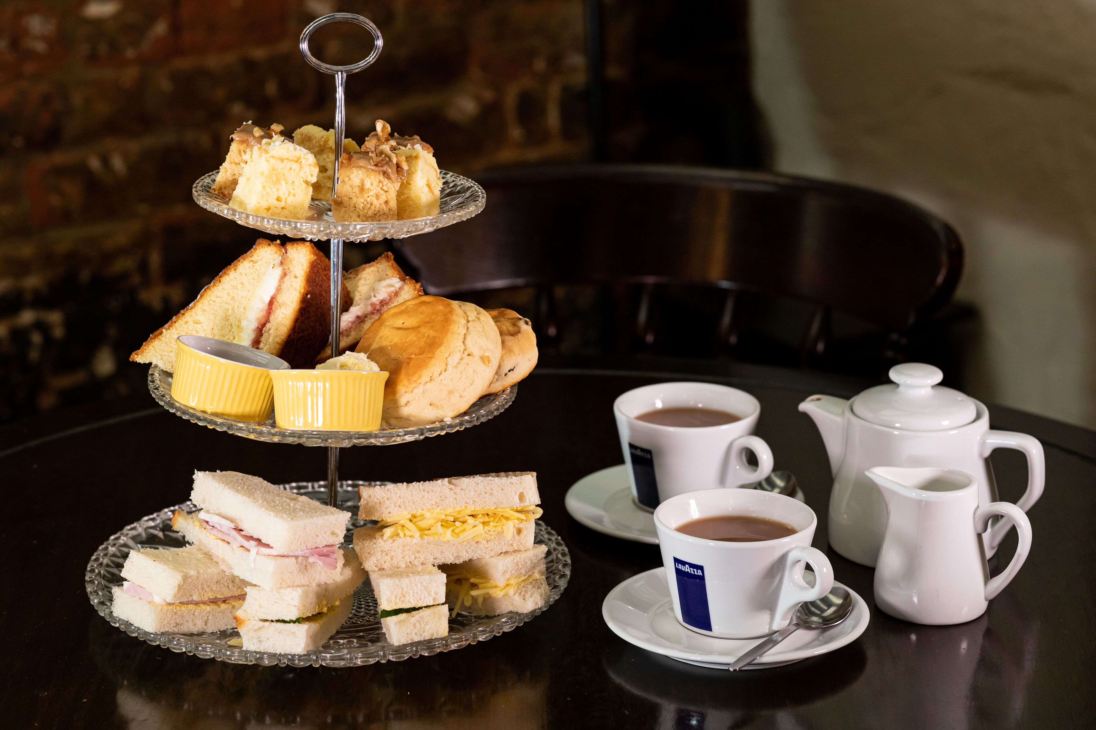 A three-tiered dessert stand with sandwiches and cakes, two cups of coffee, and a teapot on a table.