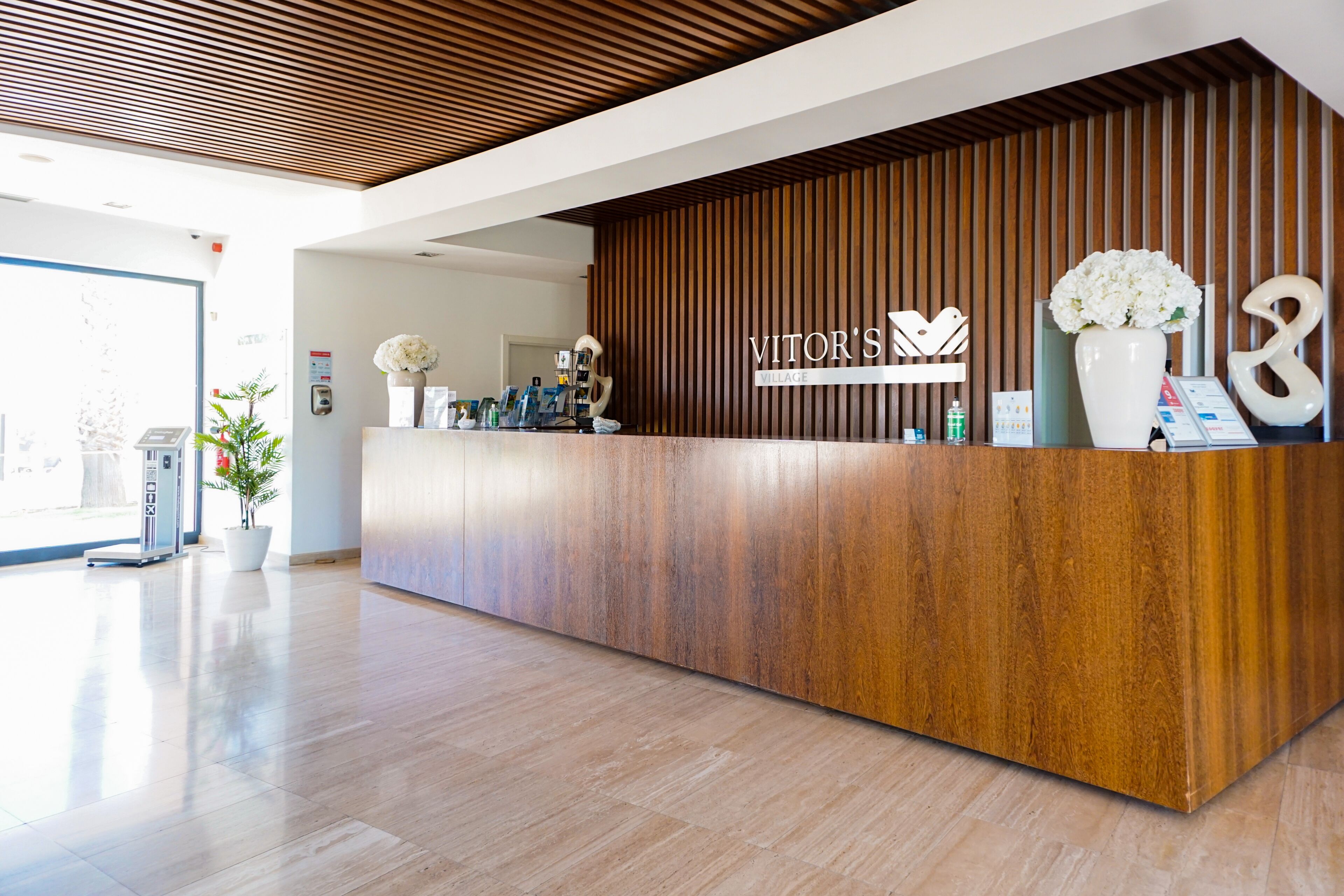 A reception desk with a wooden finish, a sign reading "VITOR'S VILLAGE," and a vase with white flowers.