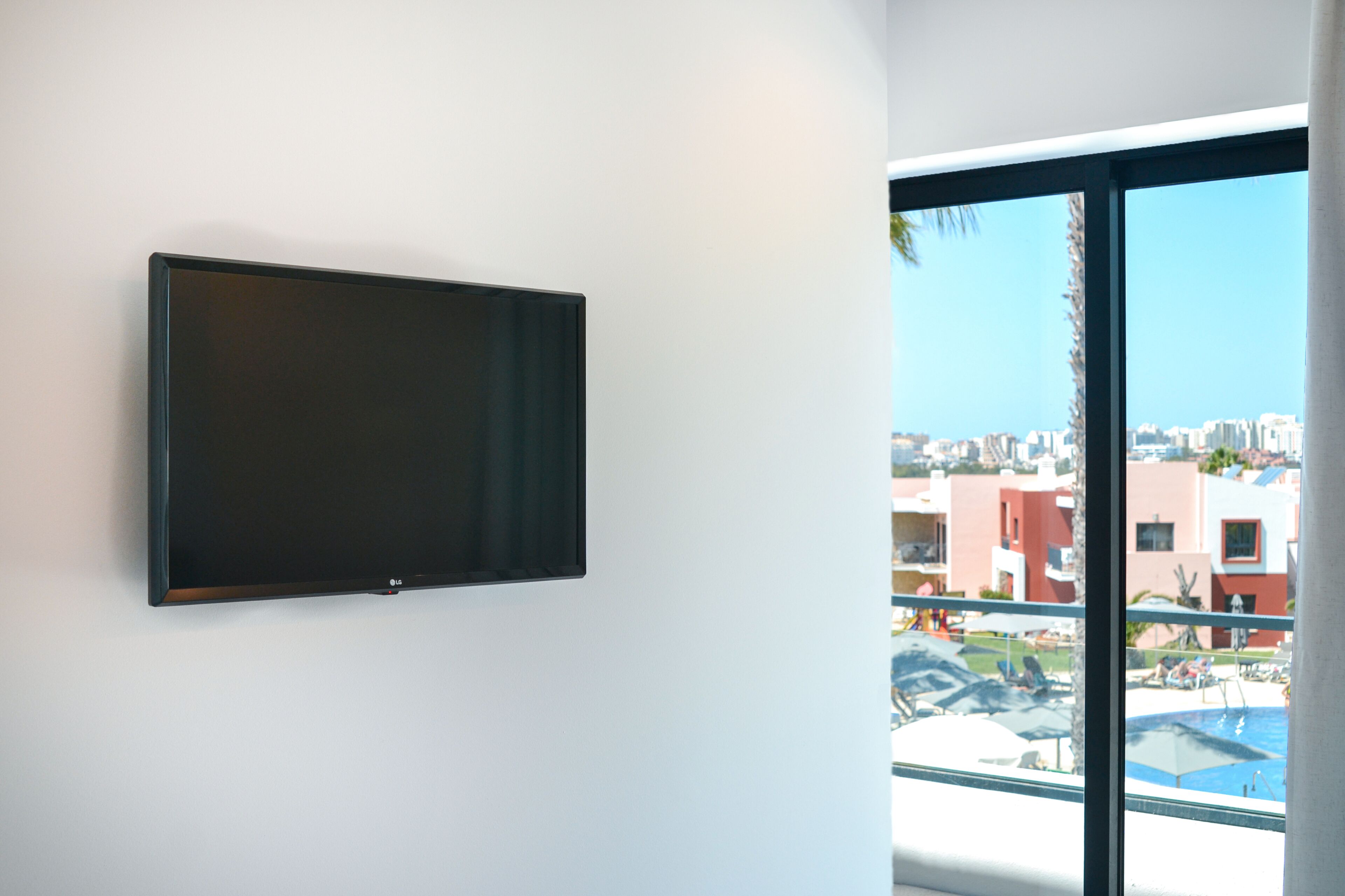Una habitación moderna con un televisor de pantalla plana montado en una pared blanca, un ventanal con vistas a una piscina y edificios.