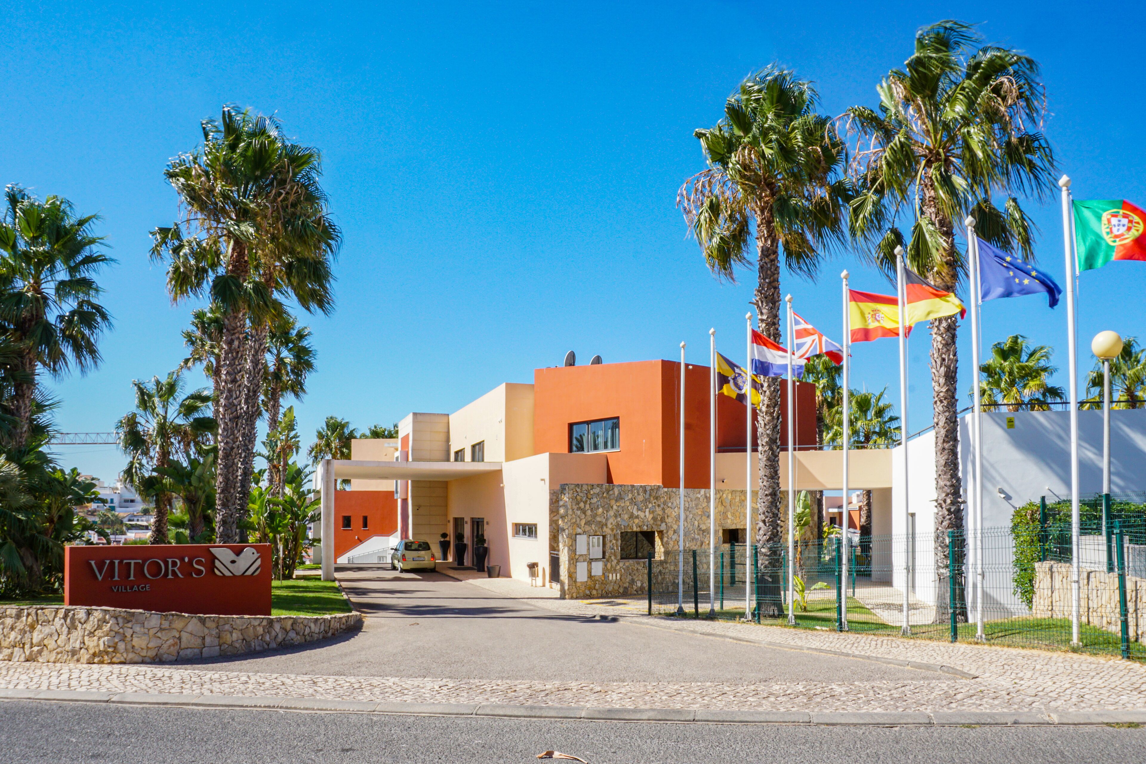 A modern building with a sign reading "Vitor's Village" and multiple flags, including the Portuguese flag, in front.