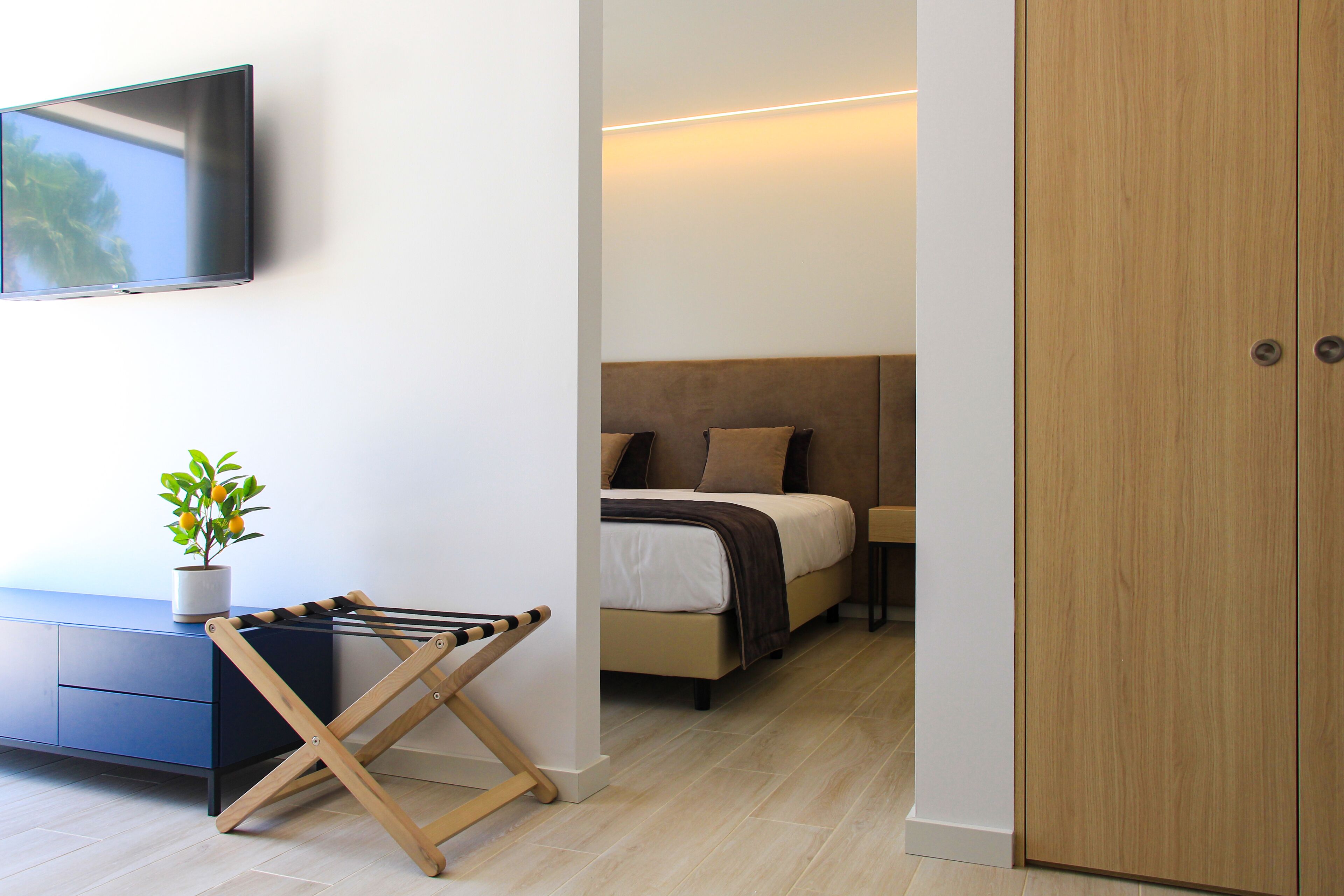 A modern bedroom with a bed, a TV mounted on the wall, a wooden door, and a small plant on a cabinet.