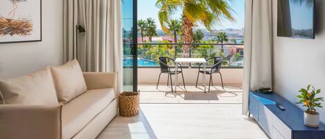 A modern living room with a sofa, a dining area with chairs, a TV, and a view of a pool and palm trees.