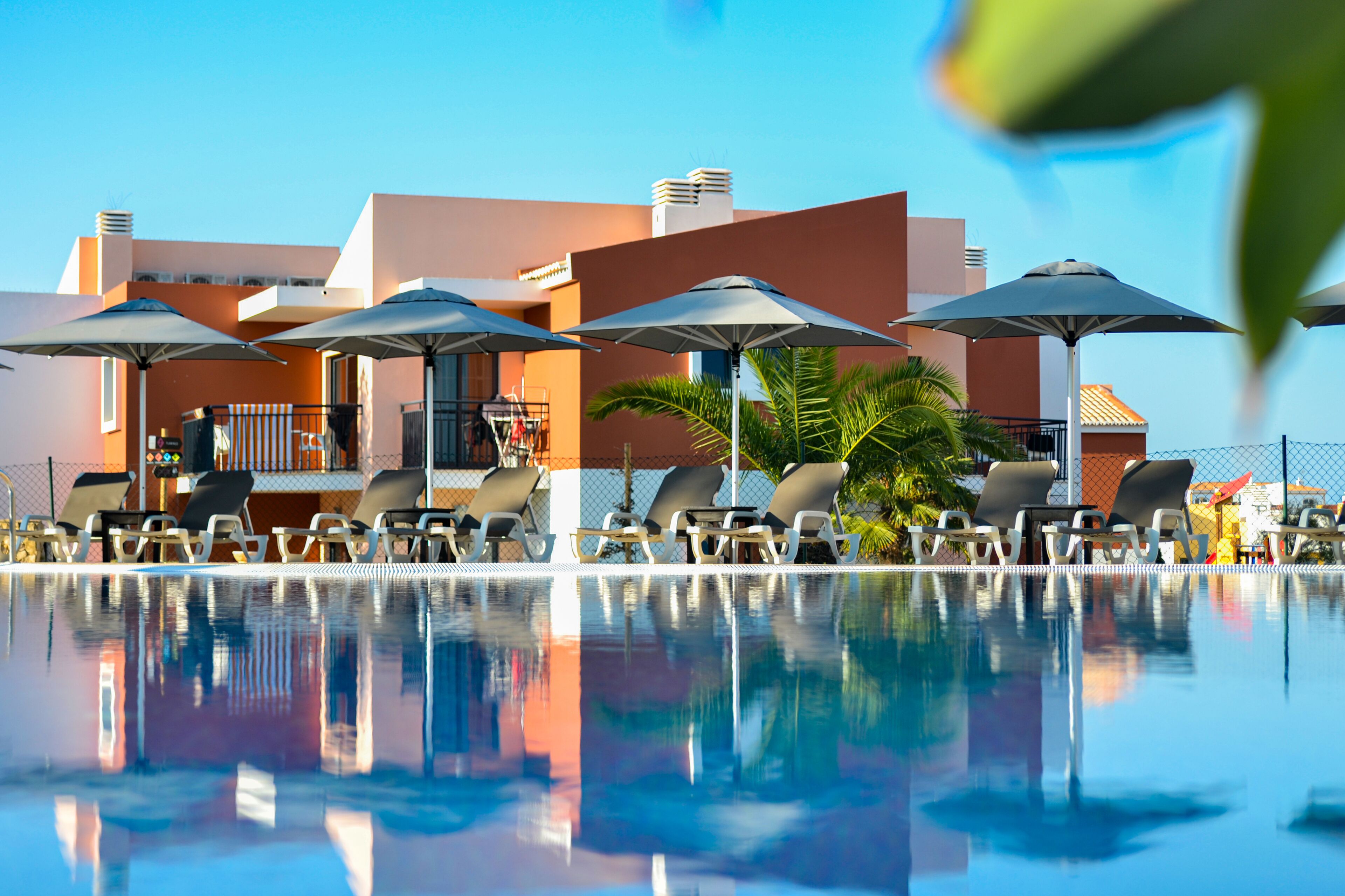 A poolside area with lounge chairs and umbrellas, reflecting the building and palm trees.