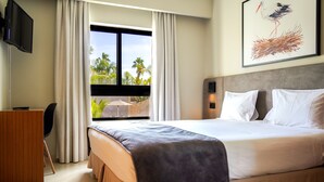 Quarto de hotel com cama, escrivaninha, cadeira, televisão e janela com vista para palmeiras.