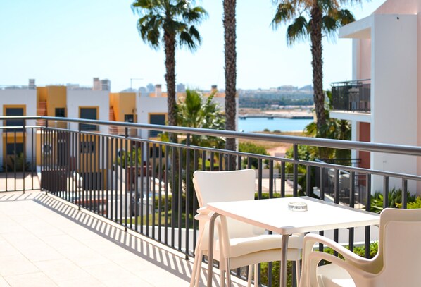 Un balcón con una mesa y sillas blancas, con vistas a una zona costera con palmeras y edificios.