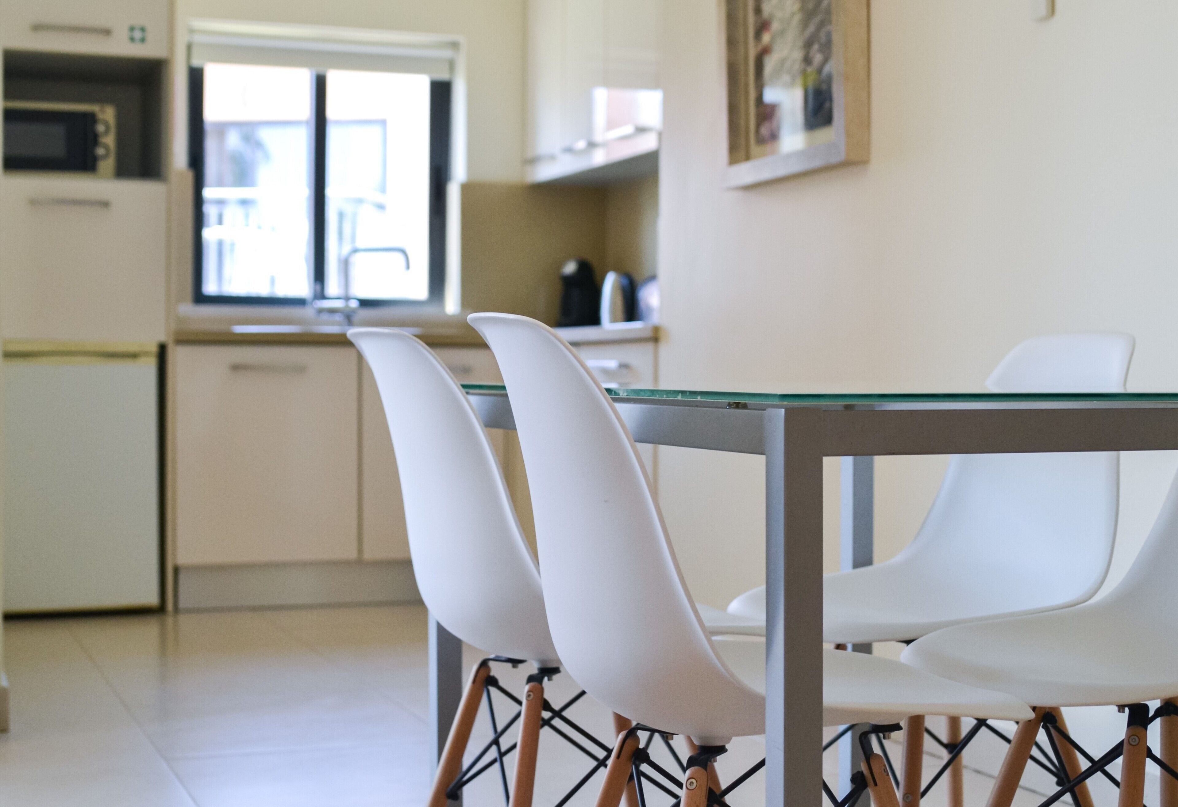 A modern kitchen with white cabinets, a microwave, and a window. There is a dining table with four white chairs.