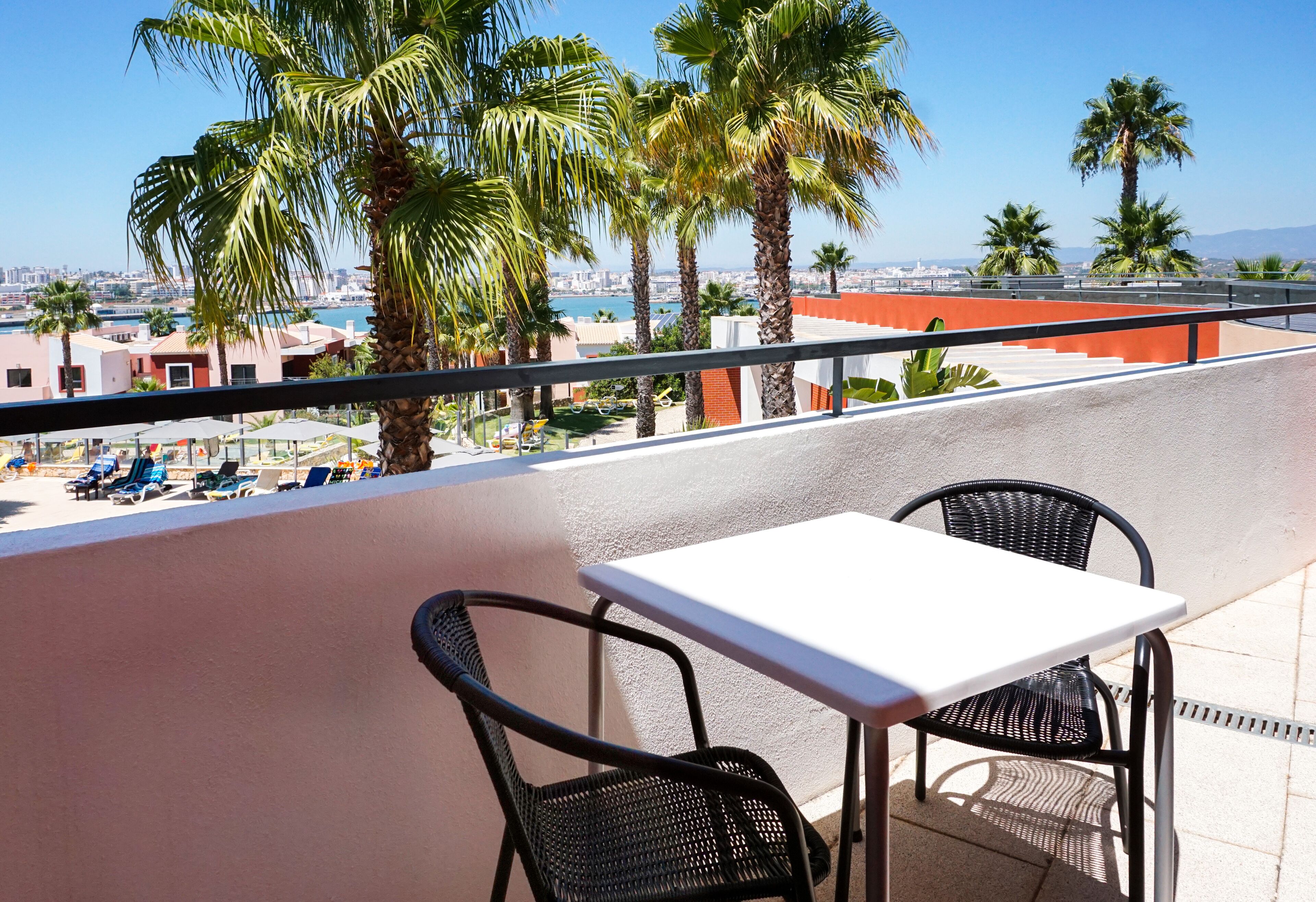 Un balcón con una mesa blanca y dos sillas, con vistas a un paisaje urbano con palmeras y al mar.