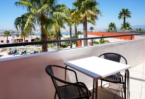 Un balcón con una mesa blanca y dos sillas, con vistas a un paisaje urbano con palmeras y al mar.