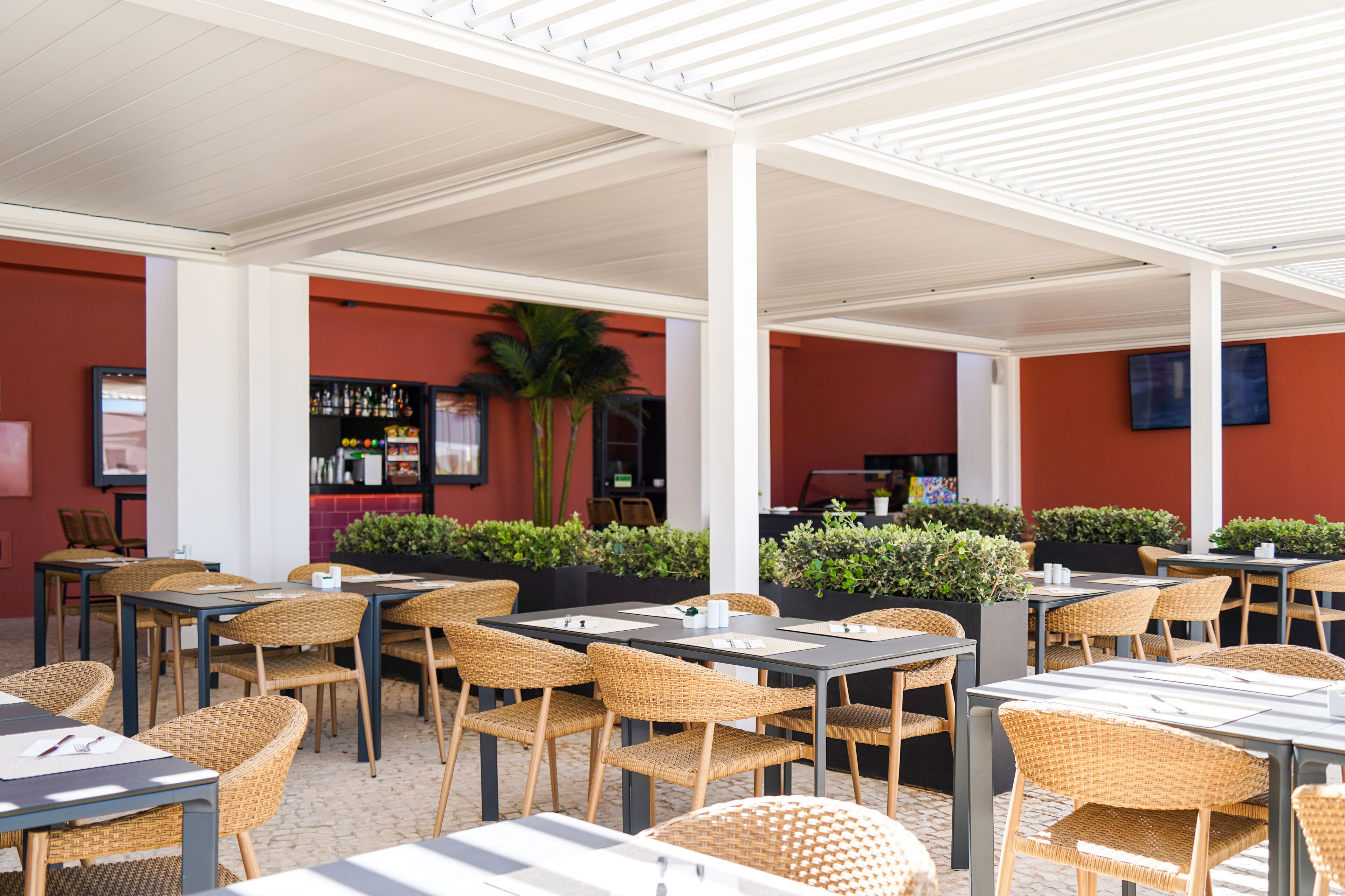 Café al aire libre con mesas y sillas de mimbre, zona de bar con estantes llenos de botellas y plantas en macetas.
