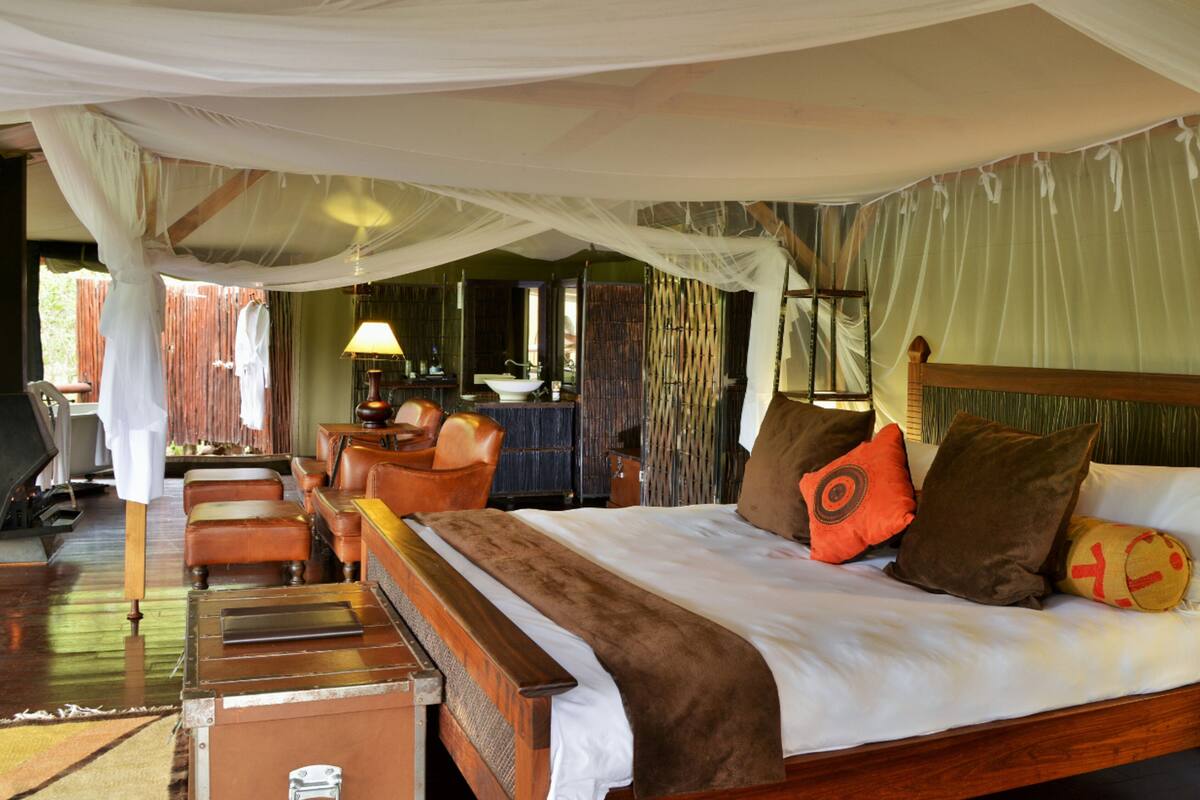 A spacious bedroom with a large bed, wooden headboard, and a canopy bed. There is a sitting area with a table and chairs, and a view of the outdoors through large windows.
