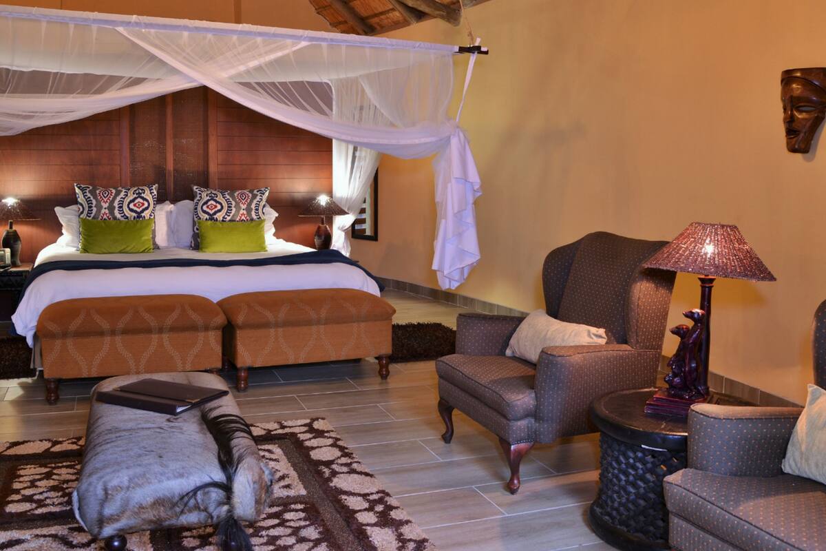 A bedroom with a canopy bed, two bedside tables, a chair, a sofa, and a small table with a lamp.