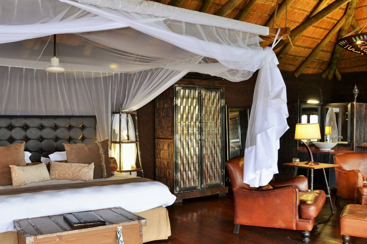 A spacious bedroom with a large bed, a wooden headboard, and a canopy bed. There is a leather armchair and a small table with a lamp.
