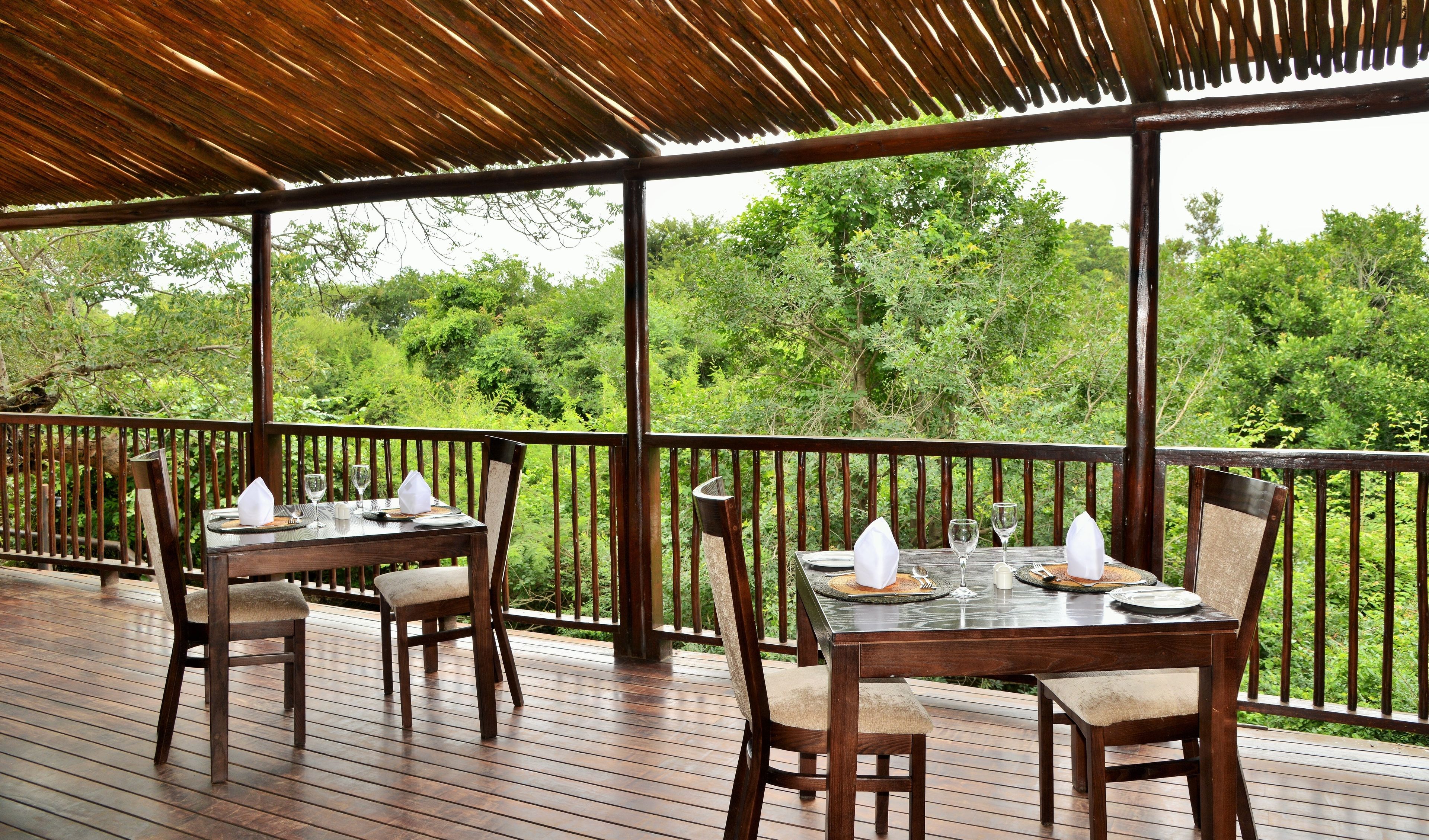 Un área de comedor al aire libre con mesas y sillas de madera, rodeada por una barandilla de madera, con vista a un bosque verde y exuberante.