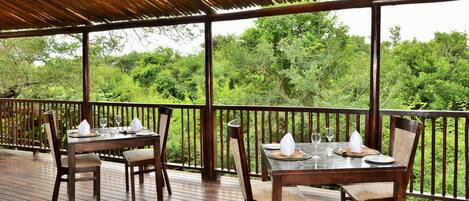 Un área de comedor al aire libre con mesas y sillas de madera, rodeada por una barandilla de madera, con vista a un bosque verde y exuberante.