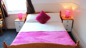 Een bed met een houten hoofdbord, witte beddengoed en een roze deken. Aan één kant staan twee nachtkastjes met lampen en een klein plankje met spullen.