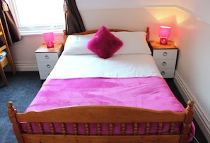 Een bed met een houten hoofdbord, witte beddengoed en een roze deken. Aan één kant staan twee nachtkastjes met lampen en een klein plankje met spullen.