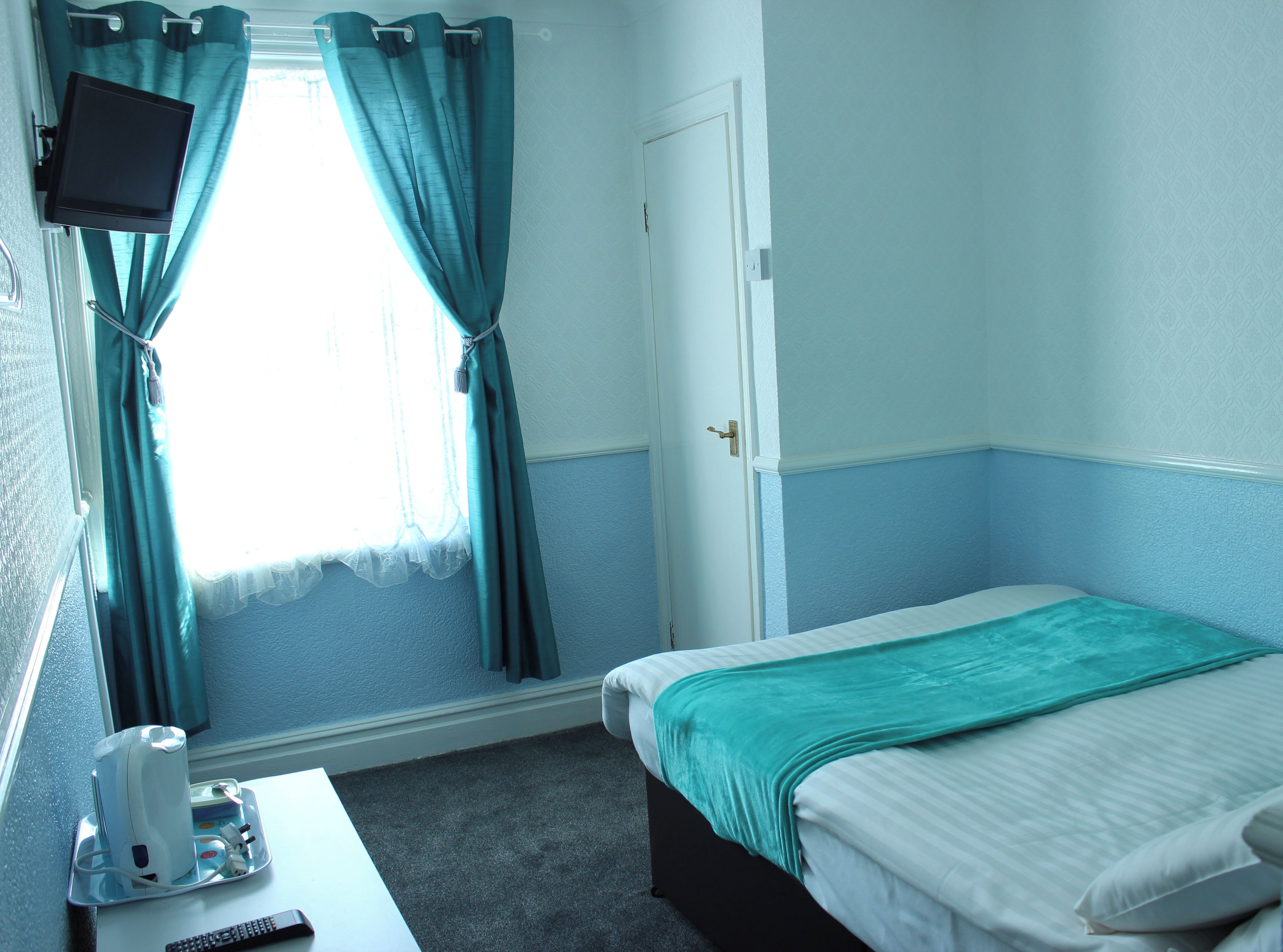 A hotel room with a bed, a desk with a remote control, a television mounted on the wall, and a window with curtains.