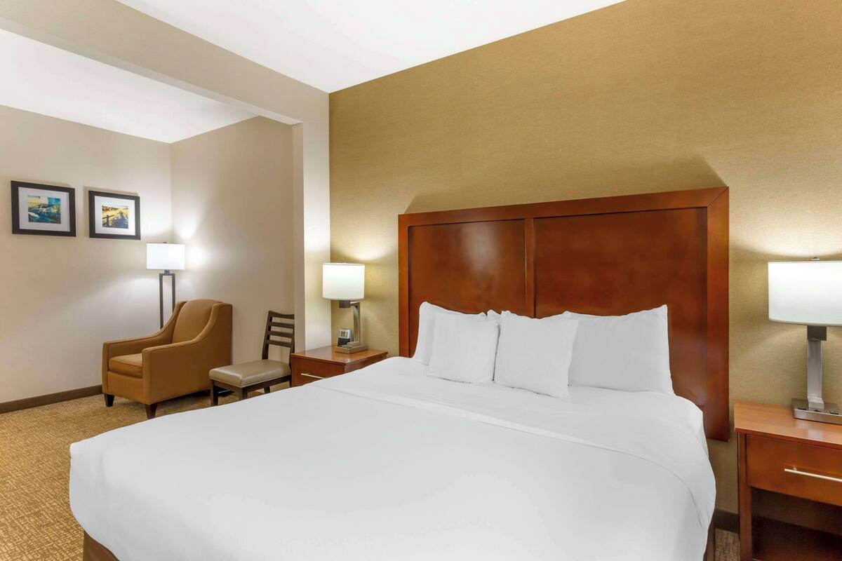 A hotel room with a large bed, a wooden headboard, a chair, a nightstand with a lamp, and two framed pictures on the wall.