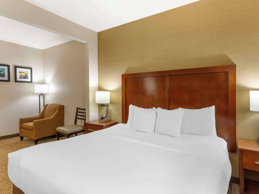 A hotel room with a large bed, a wooden headboard, a chair, a nightstand with a lamp, and two framed pictures on the wall.