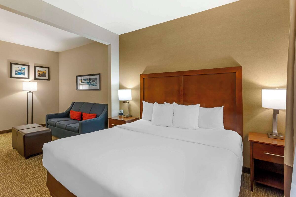 A hotel room with a large bed, a blue armchair, a small table, and a nightstand with a lamp.