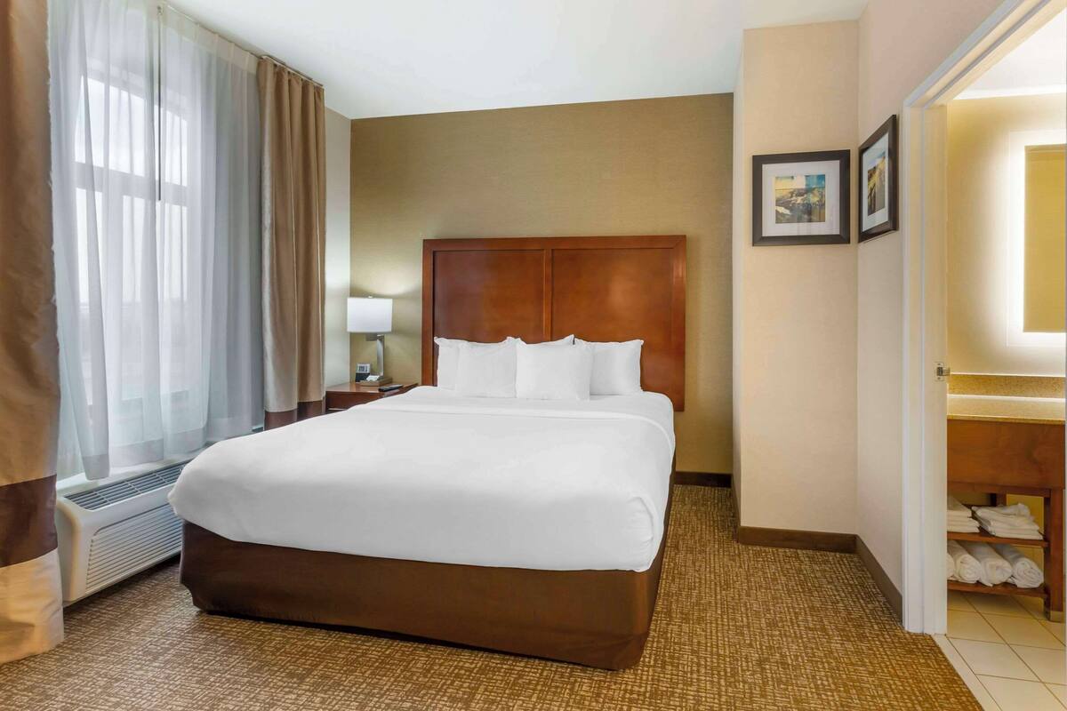 A hotel room with a large bed, a bedside table with a lamp, a wall-mounted TV, and a bathroom with a towel rack.