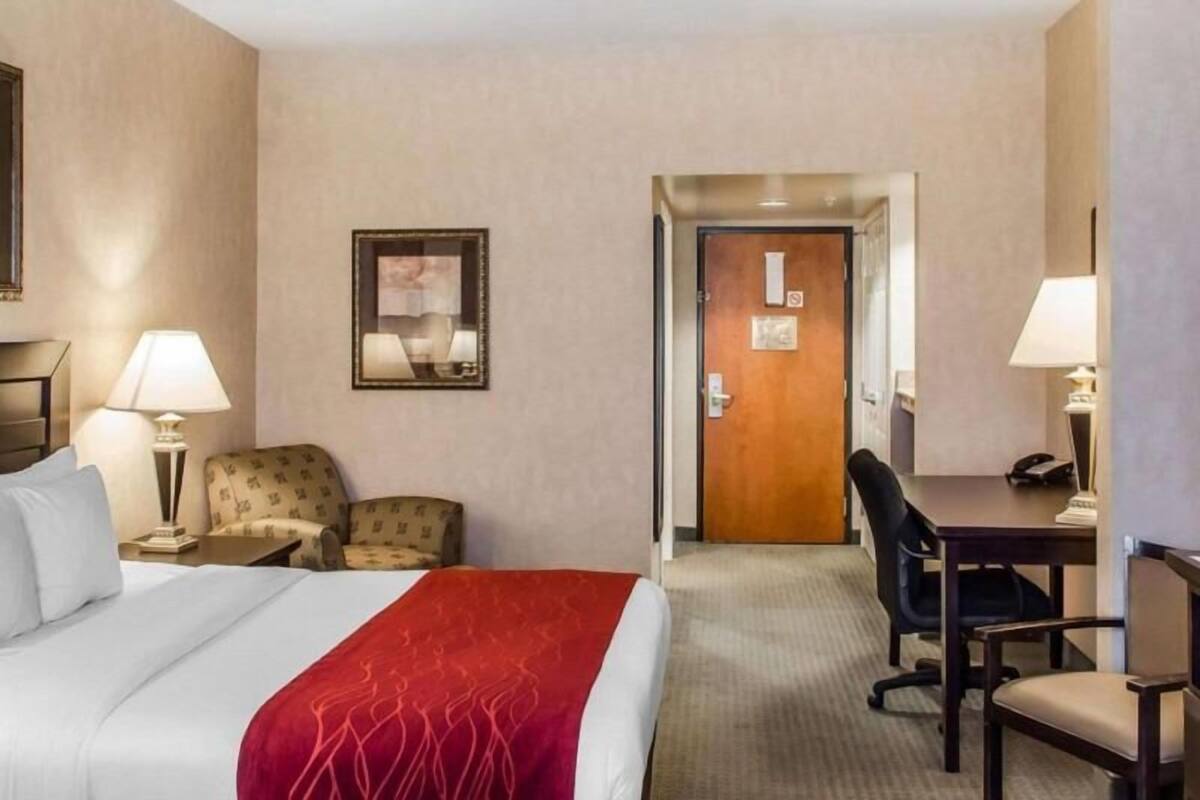 A hotel room with a bed, a desk, a chair, a lamp, and a door.
