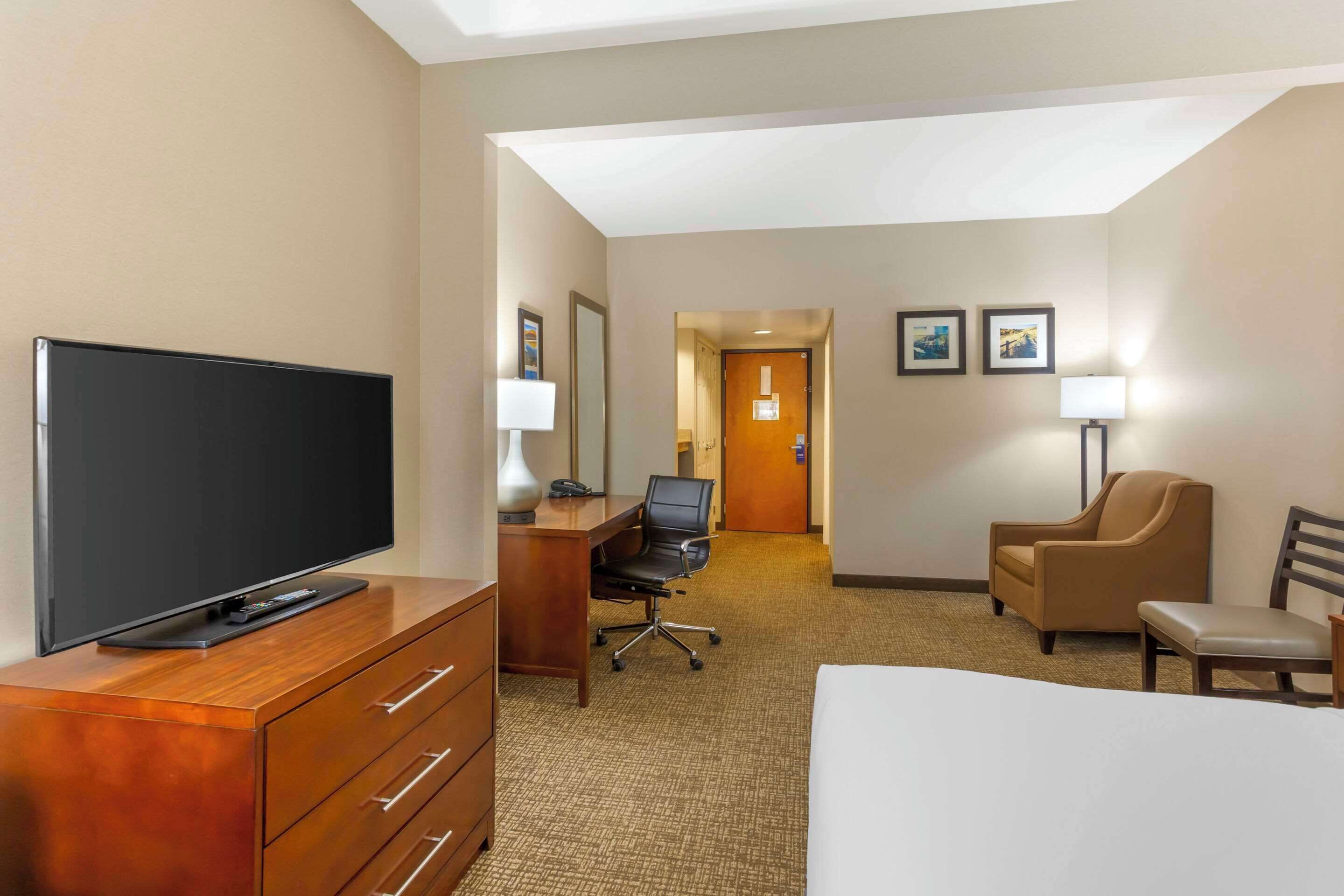 A hotel room with a flat-screen TV on a wooden dresser, a desk with a chair, a bed, and two armchairs.