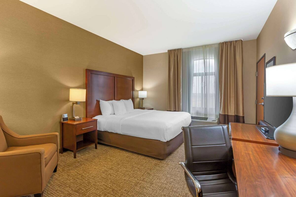A hotel room with a large bed, a desk with a chair, a desk lamp, a TV, and a window with curtains.