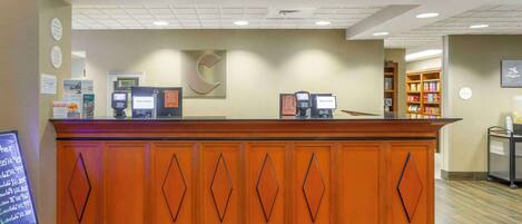A reception desk with a wooden finish and diamond-shaped paneling. There are two receptionists' stations with computers and a 'Welcome' sign above them. A signboard with room rates is visible to the left.