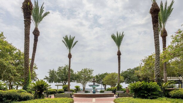 Un parque con palmeras, una fuente, bancos y vistas al mar.