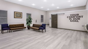 Un área de espera con un sofá marrón, dos sillones azules, una planta en maceta y una pared con una cita motivacional.