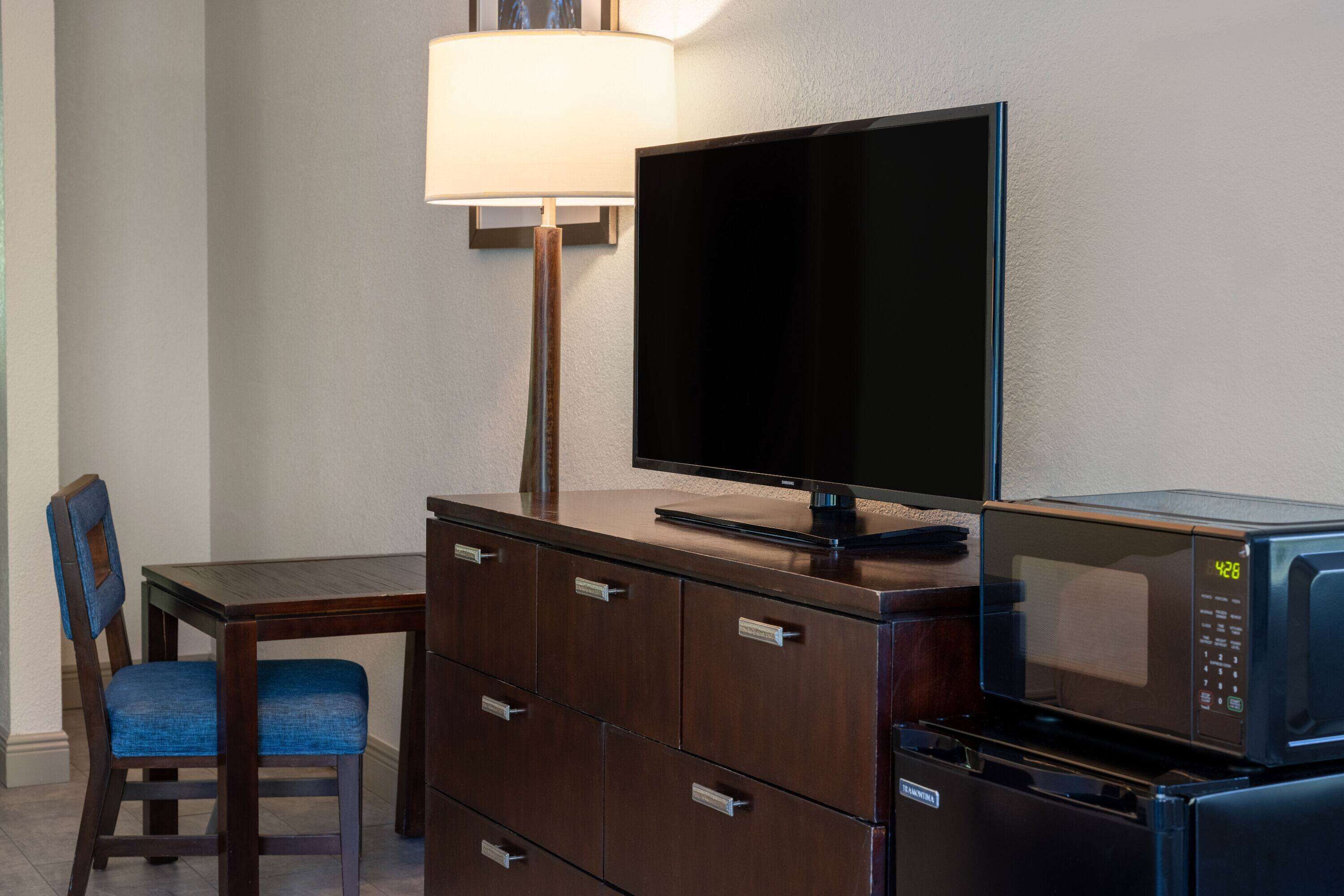 Ett hotellrum med en byrå i trä, en tv, en mikrovågsugn, en stol och en lampa.