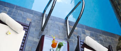 Un área junto a la piscina con dos sillones de descanso, una mesa de vidrio con una bebida y un plato de comida, y una vista despejada de la piscina.