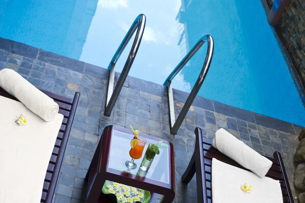A poolside area with two lounge chairs, a glass table with a drink and a plate of food, and a clear view of the pool.
