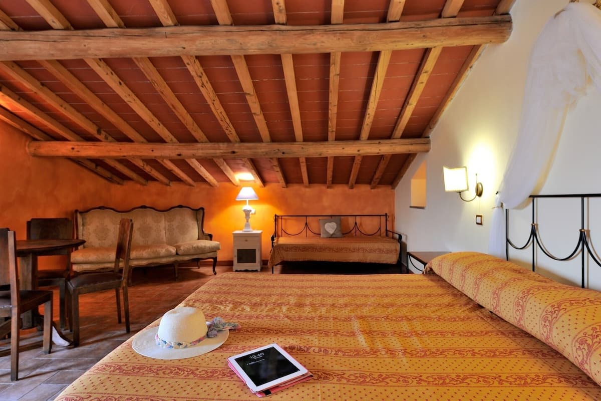 Una camera da letto mansardata accogliente, con travi a vista in legno, un letto con lenzuola a motivi decorativi, un comodino con una lampada, una sedia e un tavolino.