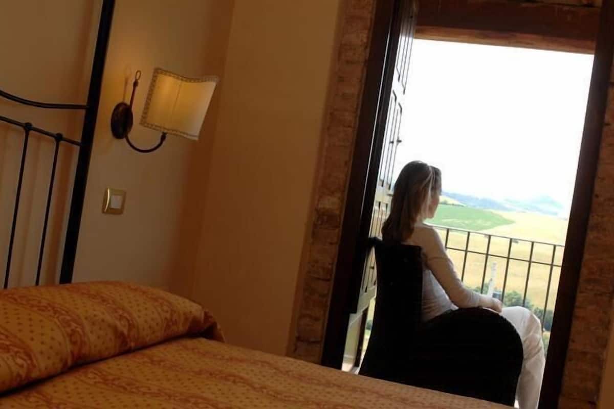 Una stanza con un letto, un comodino, una lampada e un balcone vista sulla campagna.