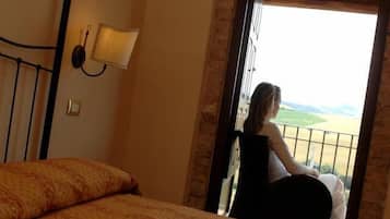Una habitación con cama, mesita de noche, lámpara y balcón con vistas al campo.