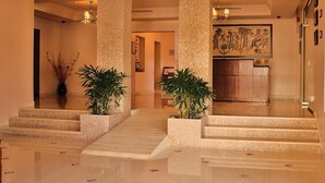 A spacious lobby with high ceilings, large columns, and a polished floor. There are two potted plants and a reception desk with a large mural on the wall. - Mosaic City Hotel (Madaba)