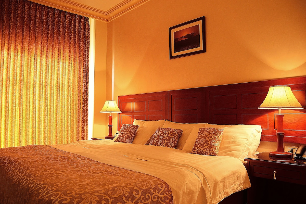 A hotel room with a large bed, two bedside lamps, a wooden headboard, and a framed picture on the wall.