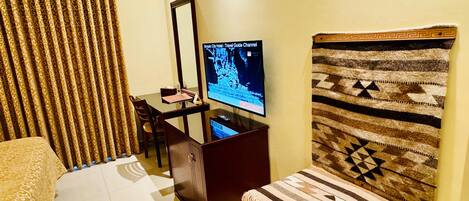 A hotel room with a bed, a desk with a computer, a television displaying a travel guide, and a patterned headboard.