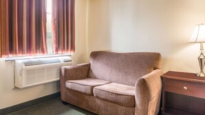 A room with a brown sofa, a lamp on a wooden side table, and a window with curtains. - Knights Inn Orillia (Orillia)