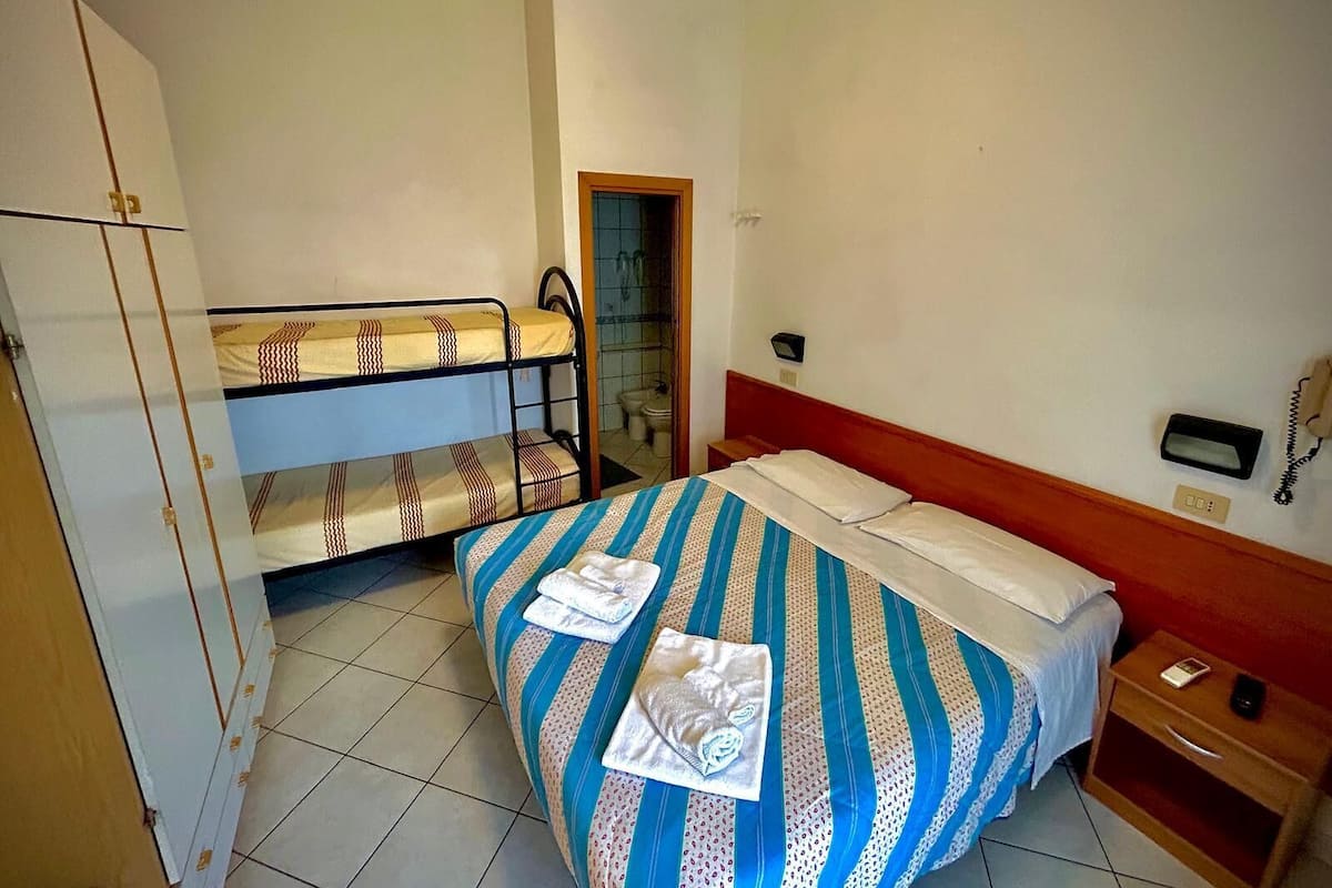 A small, compact room with a bunk bed, a single bed, a wooden wardrobe, a bedside table with a lamp, and a telephone.
