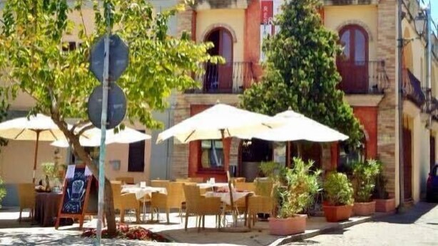 Un café al aire libre con mesas y sombrillas, rodeado de árboles y un edificio con techo de tejas.