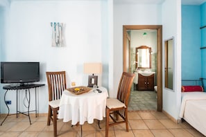 A hotel room with a TV, a small table with a lamp, two chairs, a bed, and a bathroom visible through an open door.
