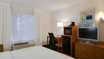 A hotel room with a bed, a desk with a chair, a television, a small refrigerator, and a wall-mounted lamp.