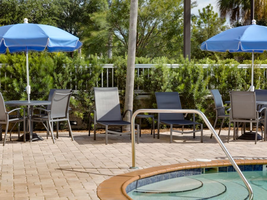 Un espace aménagé au bord de la piscine, avec des chaises longues et des tables, deux parasols bleus et une piscine dont le bord est revêtu de carreaux.